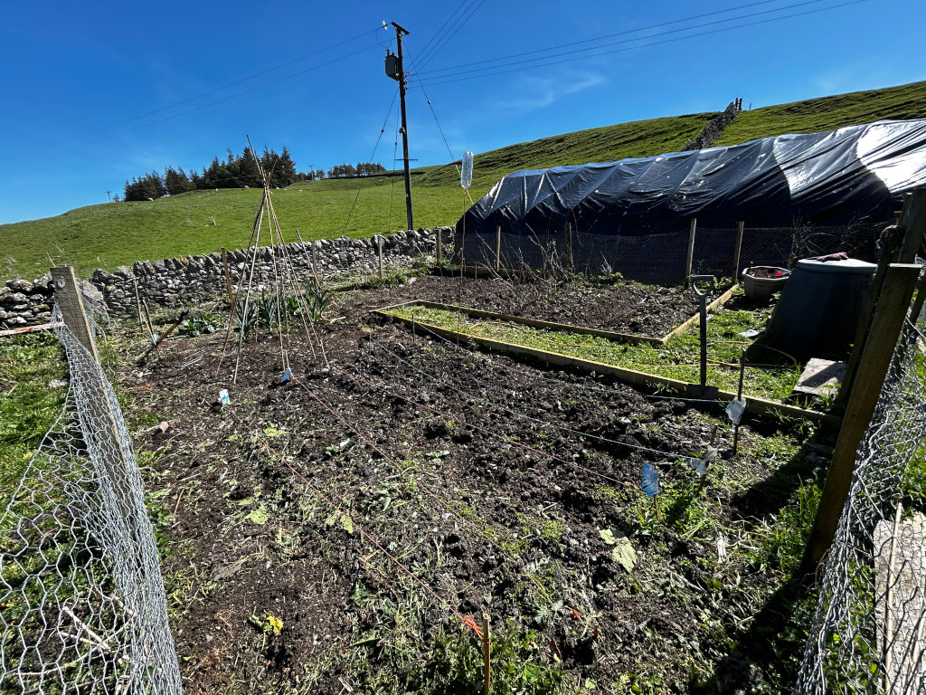 Small vegetable garden situated on a hillside. The garden is enclosed by wire fencing and features several raised beds, one of which is a defined rectangular area. There are also some wigwam structures for climbing plants, and the soil appears recently tilled. The overall setting is rural and suggests a focus on self-sufficiency or gardening.