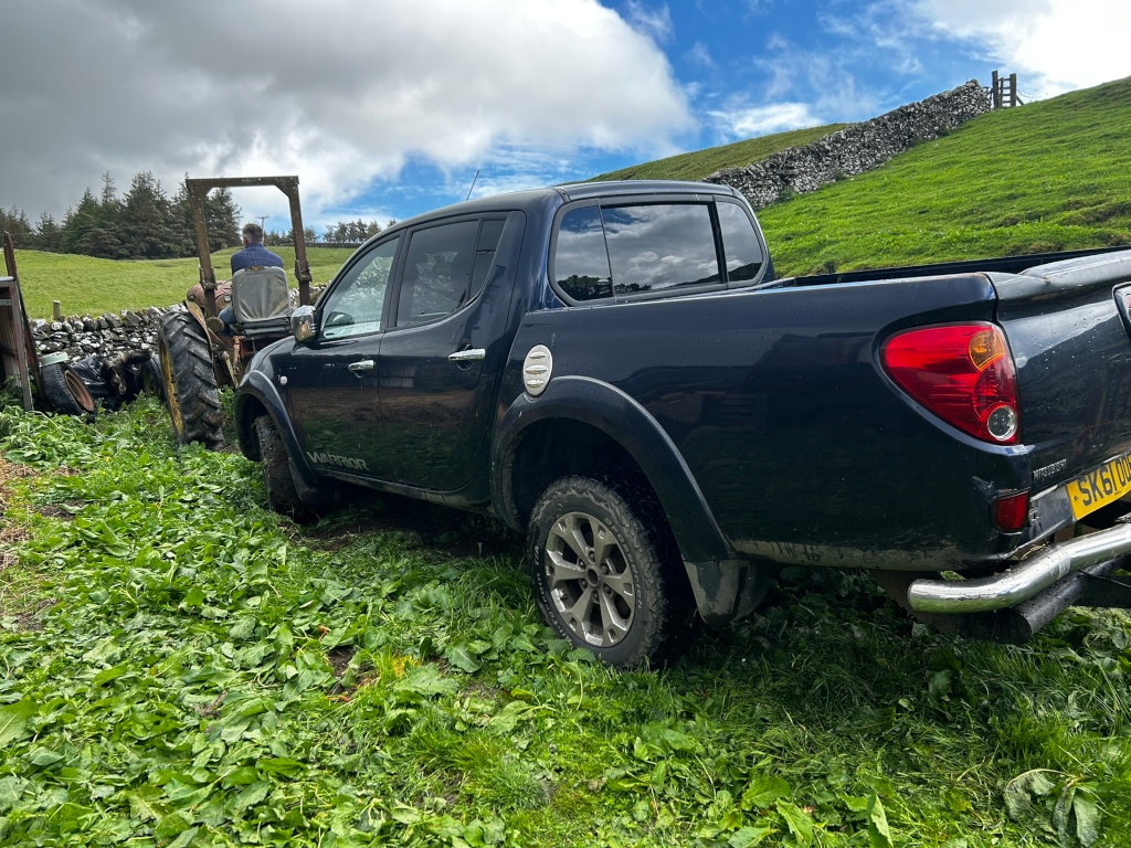 Dark-blue Mitsubishi L200 pickup truck parked on a grassy hillside next to a tractor being operated by a person. The truck is partially obstructing the view of the tractor, suggesting a working environment, possibly on a farm. The background includes a stone wall and lush green pasture under a partly cloudy sky.