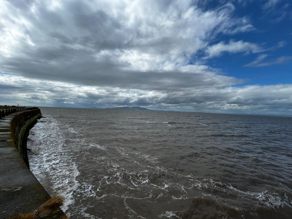 Coastal scene on a partly cloudy day. A curved concrete seawall runs along the left side of the frame, with the sea stretching to the horizon. Small waves are lapping against the wall. In the distance, a faint landmass or low-lying hills are visible against the sky. The sky is a mix of dark, heavy clouds and patches of clear blue. The overall mood is somewhat sombre yet peaceful.