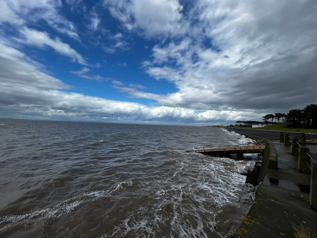 Dramatic coastal scene under a partly cloudy sky. The foreground features a partially submerged wooden ramp leading into a choppy sea. Beyond the ramp, a stone seawall extends to the right, leading toward a line of trees on a distant shore. The sky is a mix of bright blue and dark, brooding clouds, creating a strong contrast. The overall feeling is one of wild, dynamic nature, with the turbulent water emphasising the power of the sea.