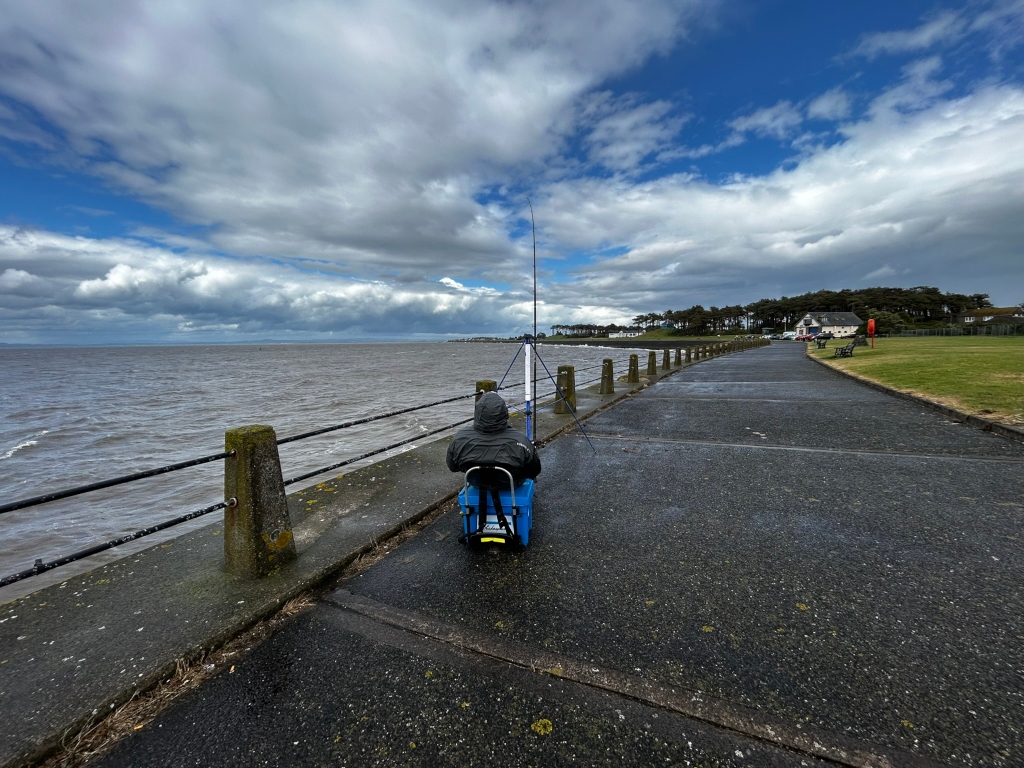 Person fishing from a seawall on a partly cloudy day. The person is seated, facing away from the camera, with their fishing rod extending out over the water. The seawall runs along a paved path that leads towards a line of trees and buildings in the distance. The sky is a mix of blue and grey clouds, suggesting a potentially changeable weather pattern.