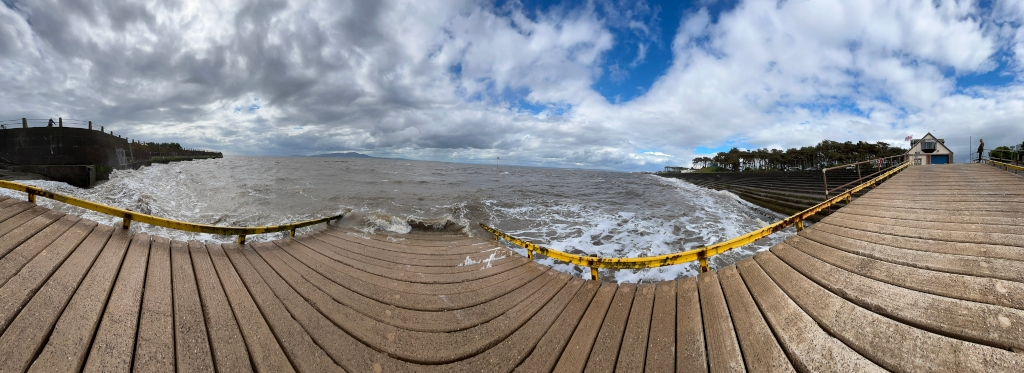 Panoramic view of a wooden pier extending into a choppy sea. Waves are breaking against the pier's edge near a seawall. In the distance, a small building is visible, situated amongst some trees. The sky is partly cloudy, with patches of blue. The overall impression is of a somewhat stormy but beautiful coastal scene.