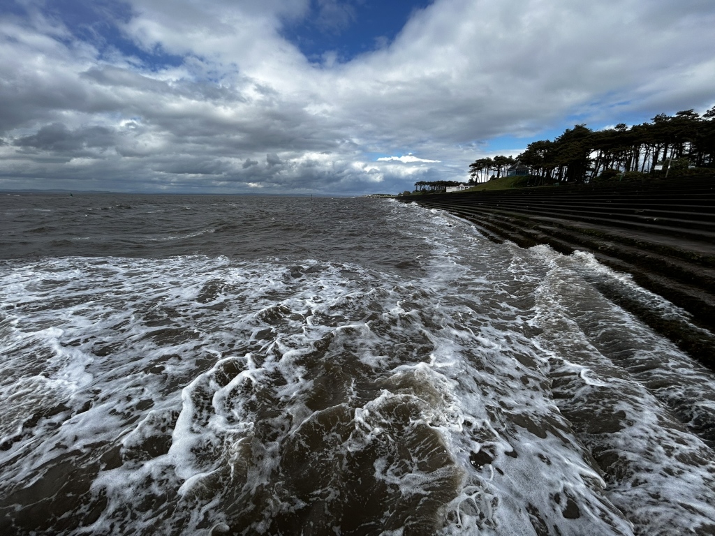 Seascape under a dramatic, cloudy sky. Dark, churning waves crash against a man-made, stepped coastal structure that runs along the shoreline, beneath a line of dark trees. The overall mood is somewhat moody and powerful, emphasising the force of the ocean against the constructed barrier.