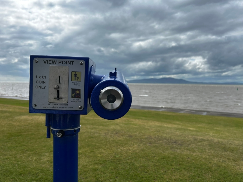 Blue coin-operated viewfinder overlooking a body of water and a distant shoreline under a cloudy sky. The viewfinder is prominently featured in the foreground, with instructions visible on its face. The background is blurred, showing the expanse of water and a hazy landscape beyond. The overall mood is somewhat subdued due to the grey sky, but the scene is peaceful.