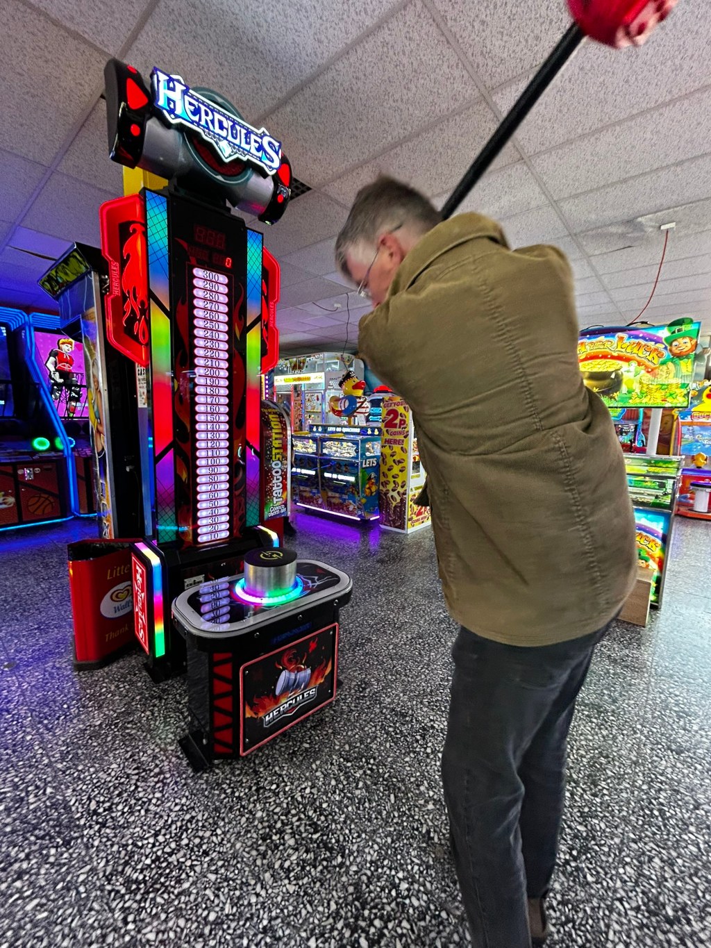 Charlie in a brown jacket playing a Hercules-branded arcade strength-testing game. The game involves hitting a target with a mallet to score points, as indicated by the illuminated scoreboard. He is in mid-swing, concentrating on the game. The setting appears to be an arcade, with various other game machines visible in the background. The overall mood is lighthearted and playful, characteristic of a recreational arcade environment