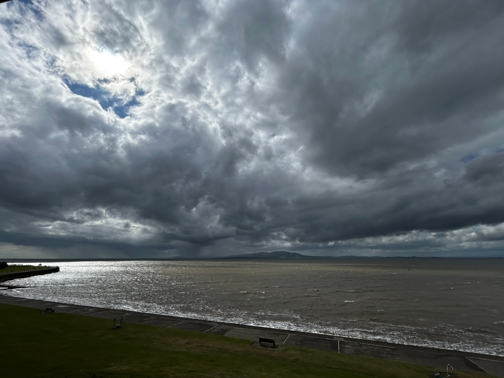 Dramatic coastal scene under a brooding, dark sky. Heavy, grey clouds dominate the upper two-thirds of the frame, with a small break revealing a sliver of brighter sky and sunlight. The lower third shows a relatively calm, dark-grey sea lapping a paved shoreline backed by a grassy area with a few park benches. A distant landmass is visible on the horizon. The overall mood is somewhat ominous and atmospheric, suggesting an impending storm or a period of intense weather.