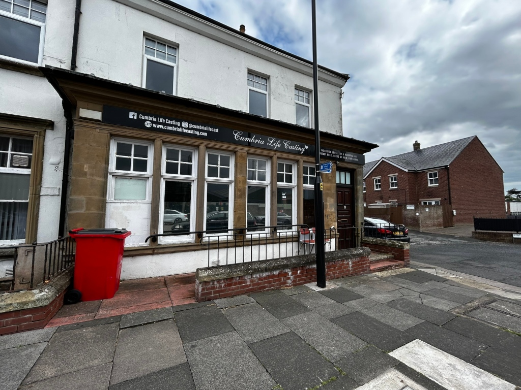 Street-level view of a storefront for Cumbria Life Casting, a business that appears to specialise in life casting.  The building is a light-colored, two-story structure with large windows. A red wheelie bin sits on the sidewalk in front of the shop.  In the background, a smaller, brick building is partially visible. The overall setting suggests a residential or commercial area in a town or city. The sky is cloudy.