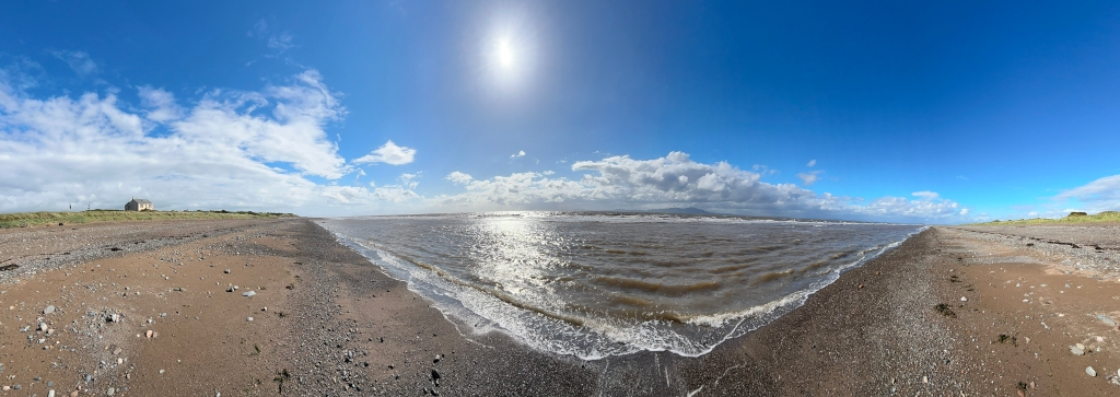 Panoramic view of a beach scene on a sunny day. The beach is composed of sand and small pebbles. The sea is relatively calm with gentle waves lapping the shore. A single, small, light-colored house is visible in the distance on the left, situated on higher ground beyond the beach. The sky is mostly clear, with some fluffy white clouds. The overall impression is one of serenity and peaceful solitude.