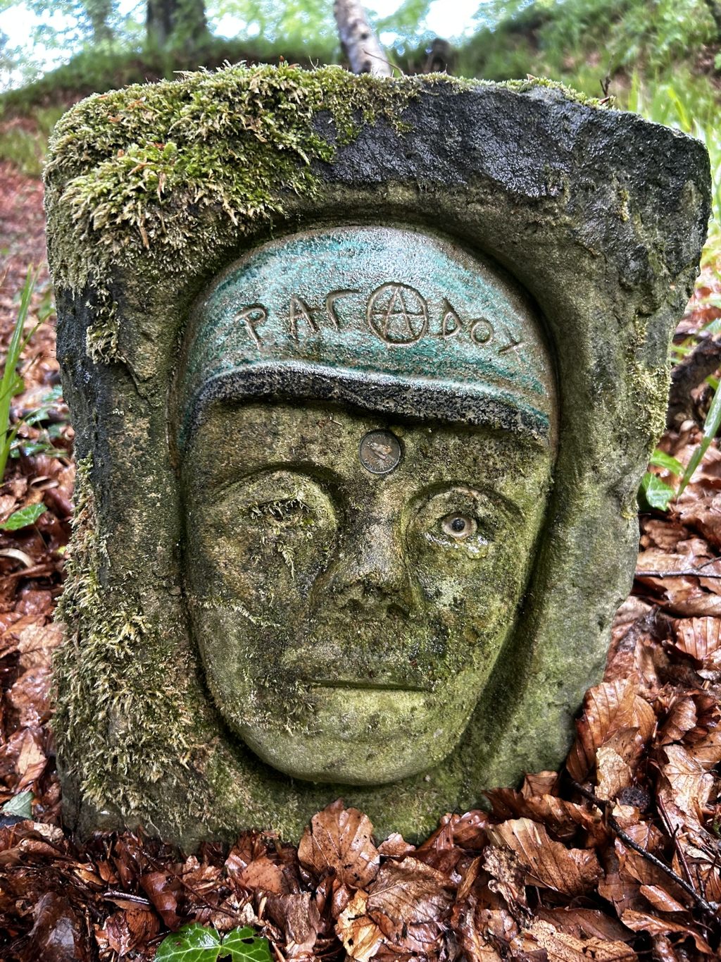 Moss-covered stone carving of a face, partially embedded in the ground amidst fallen leaves in a wooded area. The carving features a headband with an inscription and a symbol, possibly pentagram, and appears weathered and aged. The overall effect is one of ancient mystery and integration with nature.
