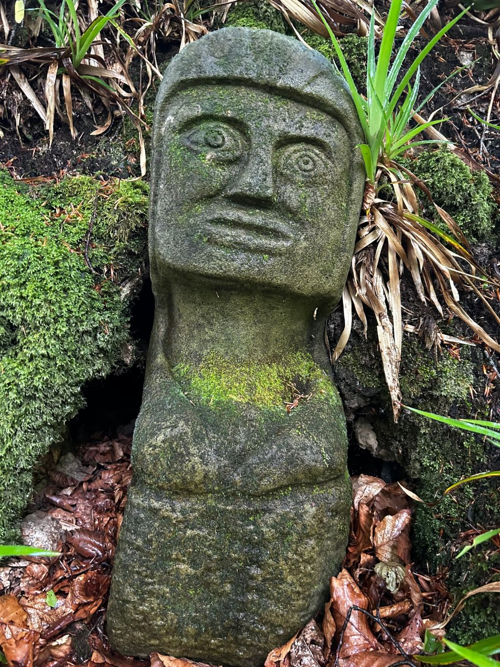 Weathered stone statue, reminiscent of a Moai statue from Easter Island, nestled amongst lush green moss and fallen brown leaves. The statue's face is carved with simple features, and its body is largely smooth. The overall impression is one of age, tranquility, and a connection to nature.