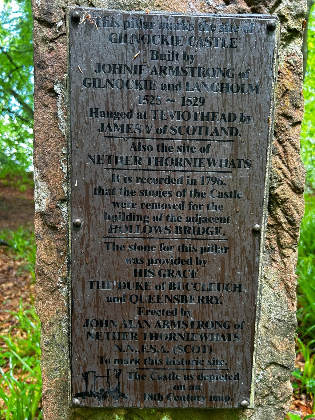 Weathered stone pillar with a metal plaque affixed to it. The plaque is an informational marker detailing the history of Gilnockie Castle. It states that the castle was built by Johnie Armstrong between 1525 and 1529, and that he was later hanged at Teviothead by James V of Scotland. The plaque further notes that the castle's stones were used to build the nearby Hollows Bridge in 1796, and that the pillar itself was erected by John Alan Armstrong to mark the historic site. The plaque also credits the Duke of Buccleuch and Queensberry for providing the stone for the pillar. A small depiction of the castle from an 18th-century map is included at the bottom. 
