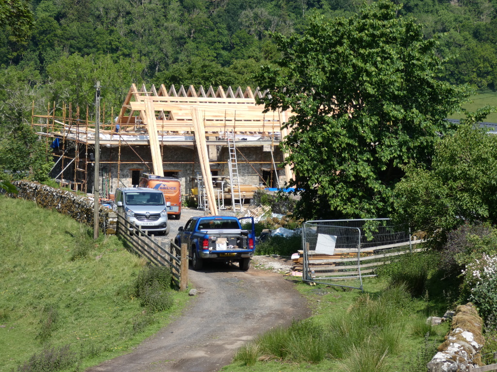 Rural construction site. A stone building is under renovation or extension, with wooden roof trusses visible. Scaffolding surrounds the building. Two vans and a pickup truck are parked nearby. The site is located in a green, hilly area with abundant trees and vegetation. A temporary metal fence is also visible near a pile of building materials.