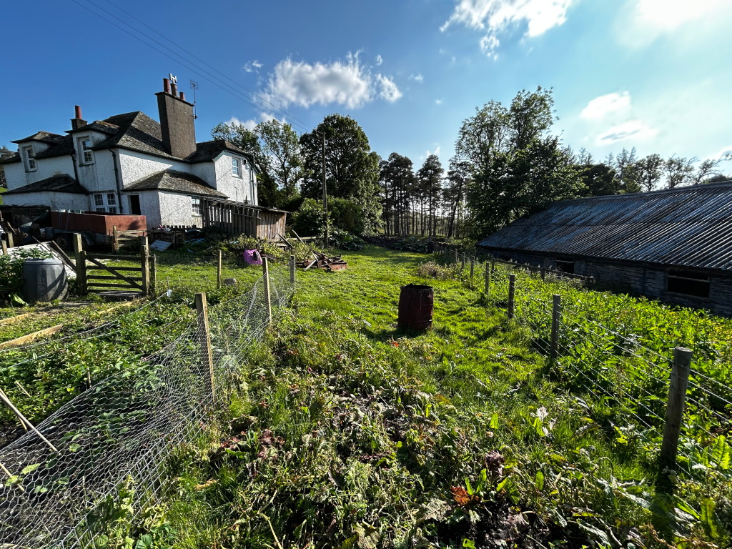 Rural scene, likely in the countryside, on a sunny day. The main focus is a grassy area, partially fenced, with a weathered stone barn on the right and a larger, whitewashed stone house on the left. The lawn shows evidence of gardening or agricultural activity.