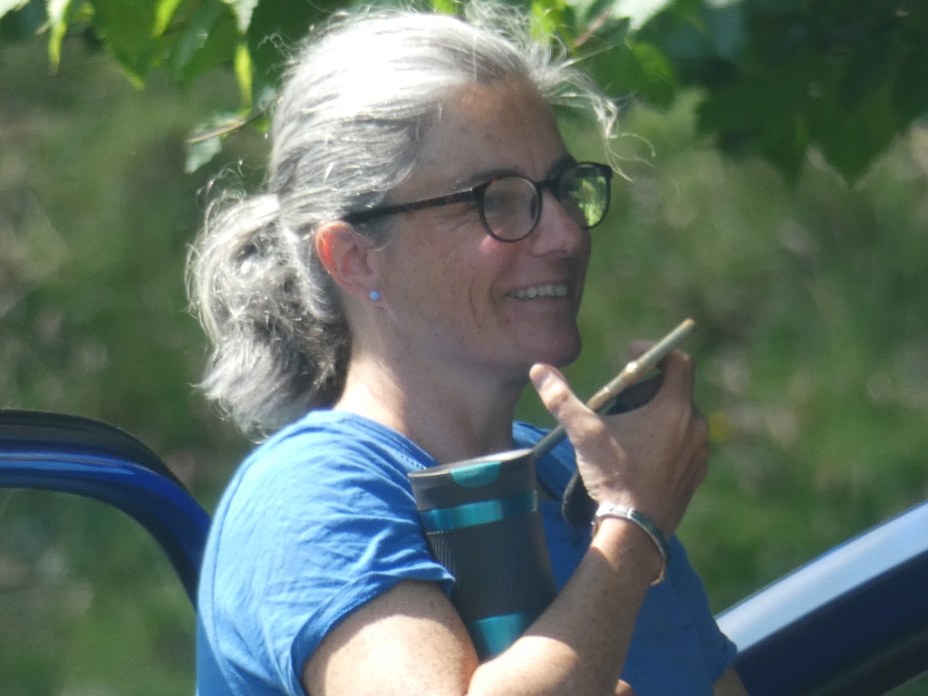 Middle-aged woman with greying hair pulled back in a ponytail. She's wearing glasses, a blue t-shirt, and a bracelet. She has a happy expression and is holding a cell phone to her ear and a travel mug under her arm. The background is blurred but shows green leafy foliage suggesting an outdoor setting possibly near some sort of vehicle. The overall impression is a casual snapshot of a woman making a phone call outdoors.