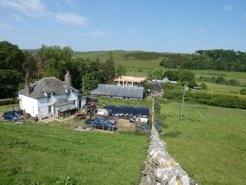 Two-story farmhouse with a slate roof. Nearby are older barns, some in apparent disrepair, and newer construction is visible, suggesting ongoing building or renovation projects. Farm equipment is scattered around the buildings. The scene is set in a green, rolling pasture with trees in the background. A dry stone wall runs diagonally across the lower right of the image.