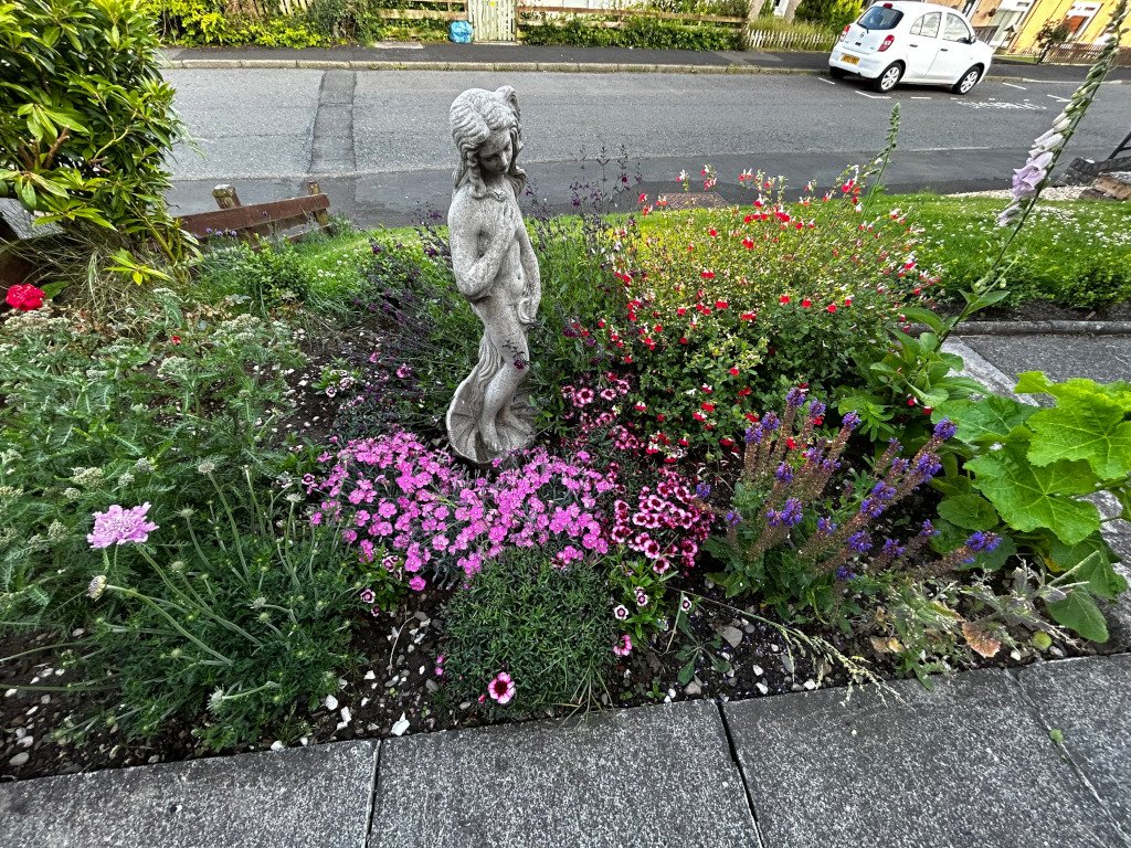 Stone statue of a woman, possibly a nymph or goddess, standing in the midst of a vibrant flower bed. The flowers include various shades of pink, red, and purple, creating a colourful and textured scene. The garden is situated along a residential street, with a portion of a house and a parked car visible in the background. The overall impression is one of peaceful beauty and careful gardening.