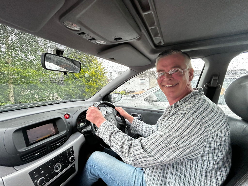 Charlie wearing glasses, he is smiling and sitting in the driver's seat of a car. He's wearing a light gray and white plaid shirt and blue jeans. It's raining outside, visible through the car windows.