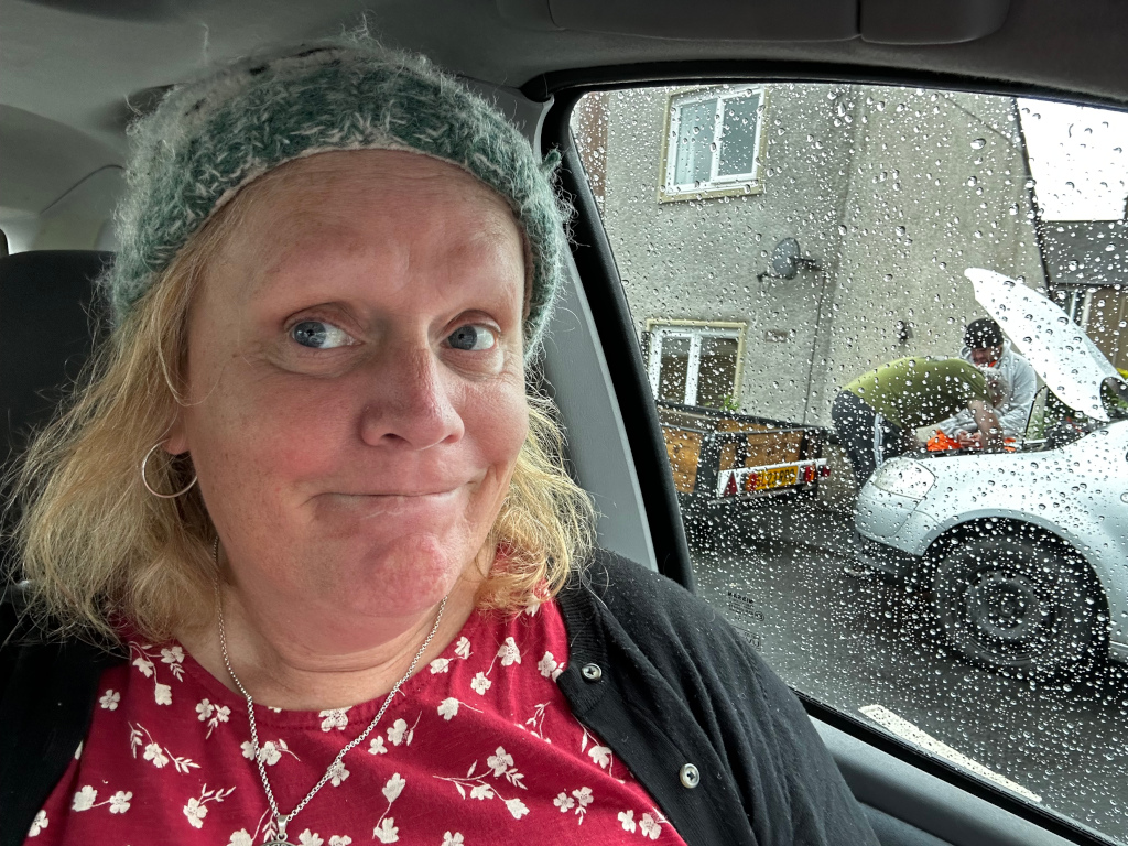 Selfie taken by Leonie inside a car. She's wearing a green knit hat and a red floral dress. Her expression is somewhat quizzical or amused. Through the rain-streaked car window, we see two men working on a car attached to a small trailer outside a house. The overall impression is of a casual snapshot, possibly capturing a moment of waiting or observation.