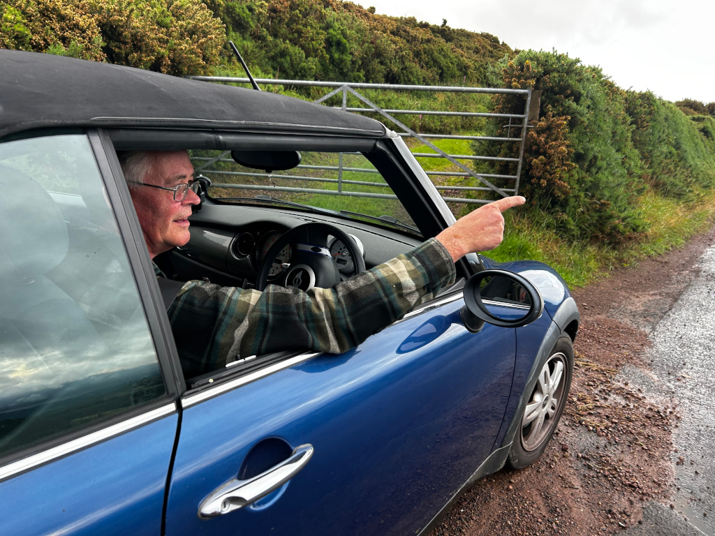 Charlie with glasses driving a blue Mini Cooper convertible. He is pointing out a gate in a hedgerow beside a country road. He appears to be taking in the scenery, perhaps pointing out a point of interest along his journey. The overall impression is one of a pleasant, rural drive.