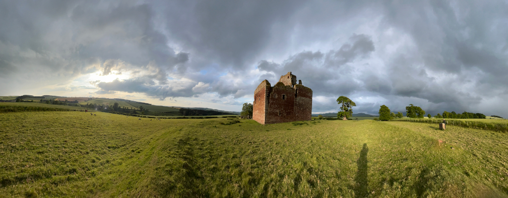 Panoramic view of a partly ruined red brick castle standing in a grassy field under a dramatic, cloudy sky. The castle is the central focus, with the field extending to the horizon where rolling hills are visible. The sky is a mix of dark, brooding clouds and lighter areas suggesting a break in the weather. A person's shadow is cast onto the grass in the foreground. The overall scene evokes a feeling of history and possibly impending weather change.