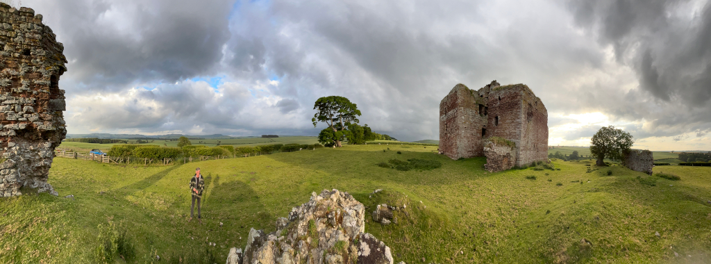 Panoramic view of the ruins of a stone castle set in a grassy field under a dramatic, cloudy sky. Charlie is standing near the ruins, providing a sense of scale. The remains of the castle are partially intact, with some walls still standing, while others are crumbled. The surrounding landscape is relatively flat, with some low-lying vegetation and a few trees.