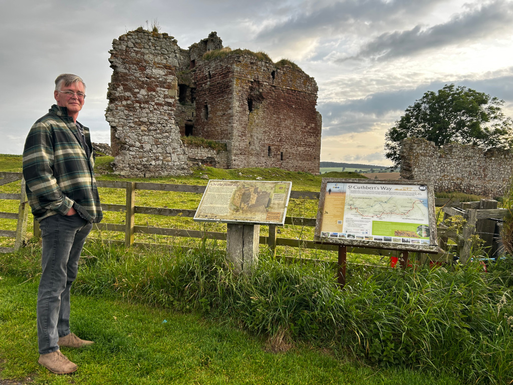 Charlie standing in front of the ruins of a stone castle. Two informational signs about the St. Cuthbert's Way are visible next to him. The setting appears to be a grassy field with a wooden fence. The overall mood is peaceful and historical.