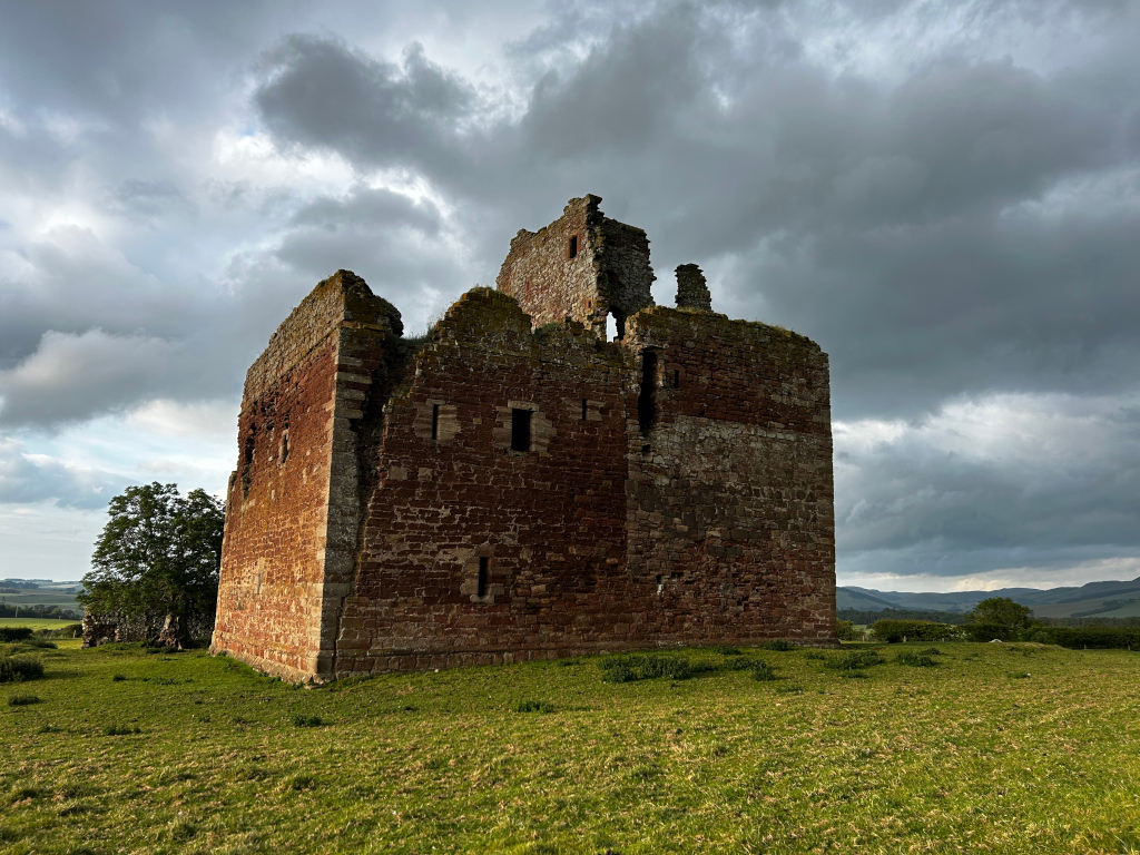 Ruins of a stone tower, possibly a castle or keep, set against a dramatic, cloudy sky. The structure is made of reddish-brown stone and is partially collapsed, with visible gaps and missing sections of wall. The surrounding landscape is green pasture. The overall mood is somewhat melancholic and evokes a sense of history and the passage of time.