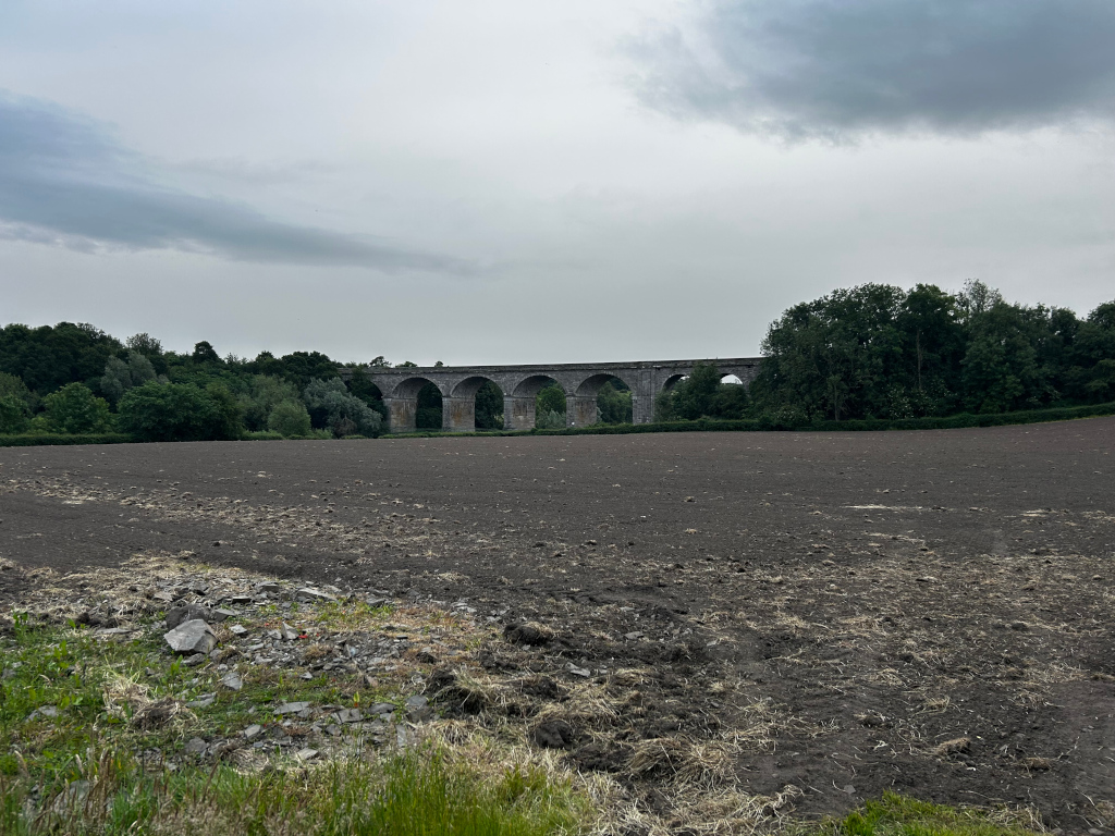 Long stone viaduct in the background, stretching across the width of the image. The viaduct is situated behind a line of trees and a large, dark brown, tilled field that dominates most of the foreground. The sky is overcast and gray. The overall impression is one of quiet, rural landscape under a sombre sky.