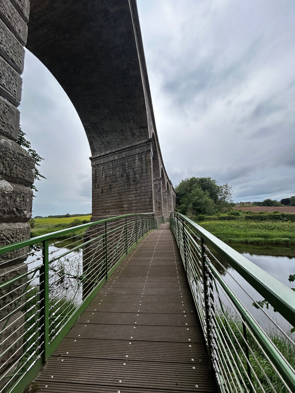 Modern pedestrian bridge extending from the viewer's perspective towards a large, old stone viaduct. The bridge has a dark brown wooden deck and green metal railings.  The viaduct arches over a calm body of water and lush green landscape. The sky is overcast. The overall impression is one of peaceful serenity and the contrast between old and new architecture.