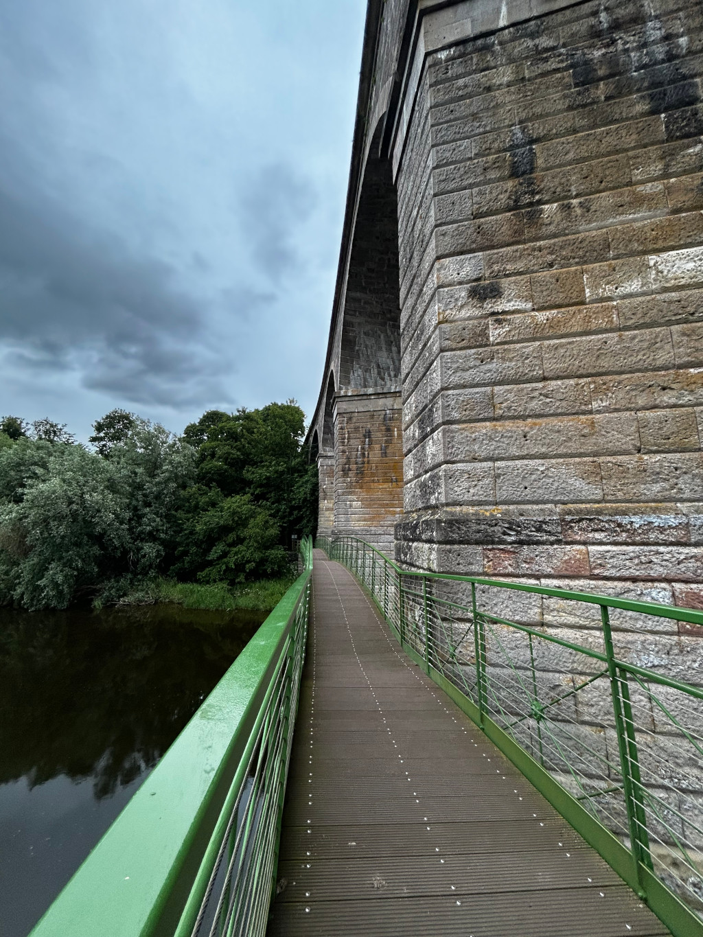Modern green metal pedestrian bridge attached to a large, old stone viaduct. The viaduct is made of large, uneven blocks of grey and brown stone. The bridge leads across the top of the viaduct and over a dark river or canal. Lush green vegetation borders the waterway. The sky is overcast, suggesting a cloudy or somewhat gloomy day. The perspective of the image is from the bridge looking towards the viaduct and river.