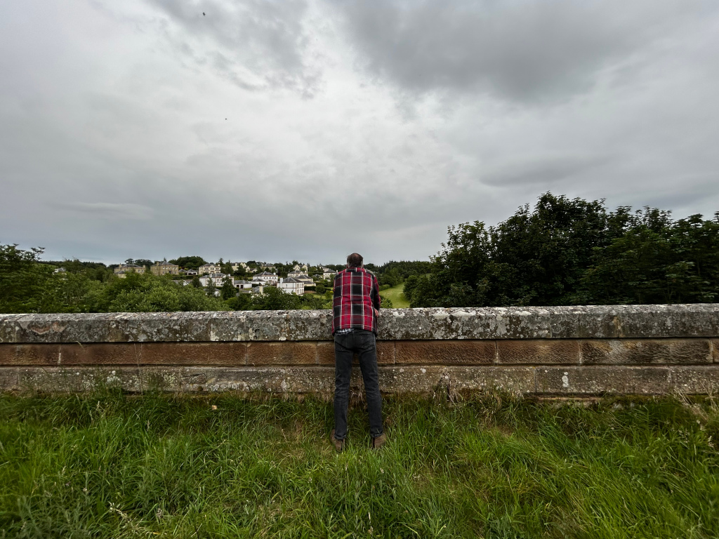 Charlie with his back to the camera, standing on a low stone wall and looking out at a small town nestled amongst green hills under a gray, overcast sky. He is wearing a red and black plaid shirt and dark pants. The overall mood is contemplative or perhaps slightly melancholic, enhanced by the muted colours and cloudy sky.