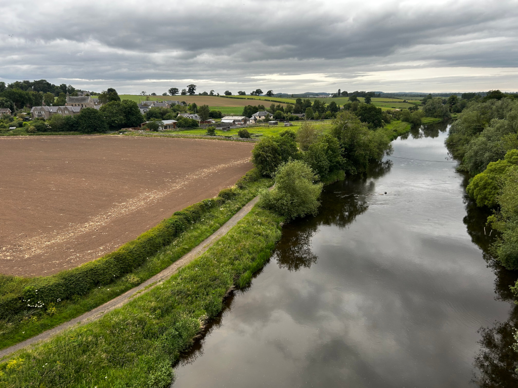 High-angle, long shot of a river meandering through a rural landscape. To one side of the river is a large, tilled field, and on the other lush green banks with trees. A narrow path runs alongside the river, separating the field from the riverbank. In the distance, a small village or collection of houses is visible. The sky is overcast, giving the scene a somewhat muted but peaceful atmosphere.