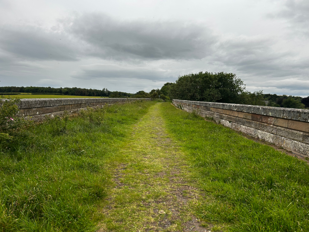 There is a grassy path going down the middle of a stone viaduct. On both sides of the path are low stone walls and tall grass. A flat, verdant landscape can be seen in the distance beneath a cloudy sky. On the whole, it looks like a peaceful and quiet place in the country.