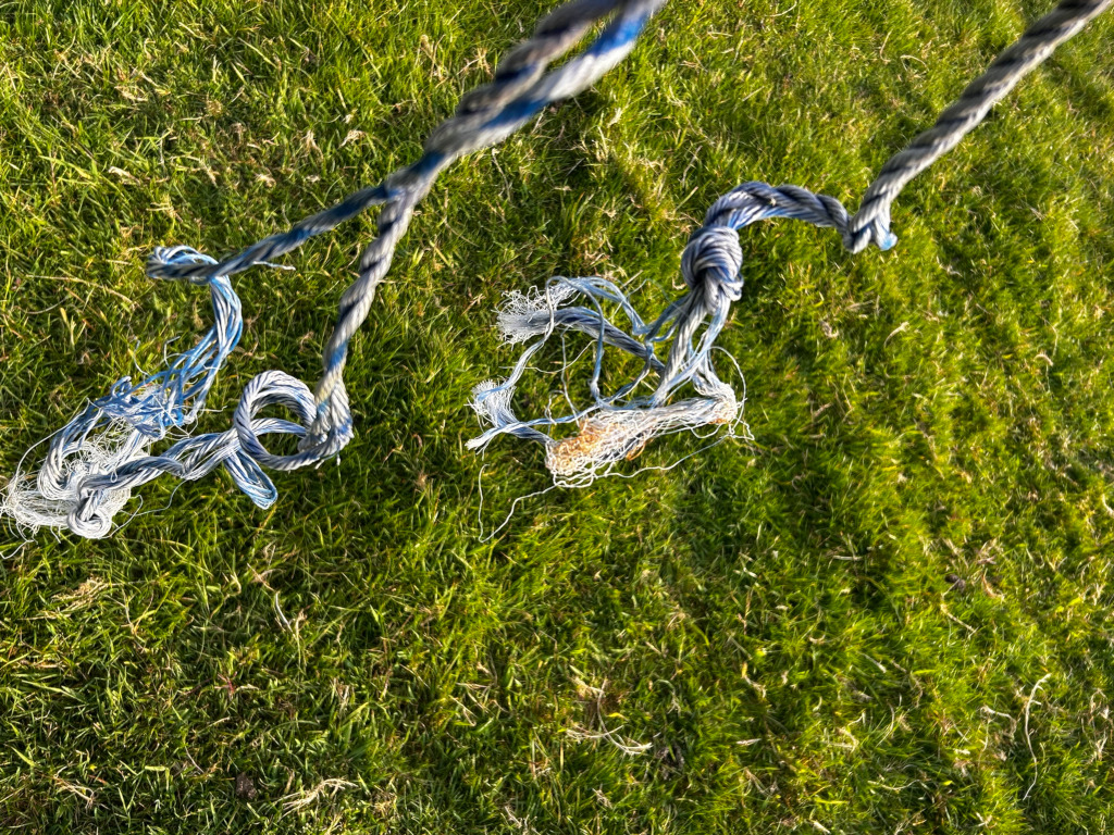 Section of frayed rope hanging down against a background of short, green grass. The rope is predominantly grey/blue with some white fraying visible, particularly at the ends. The rope appears knotted and tangled in places.