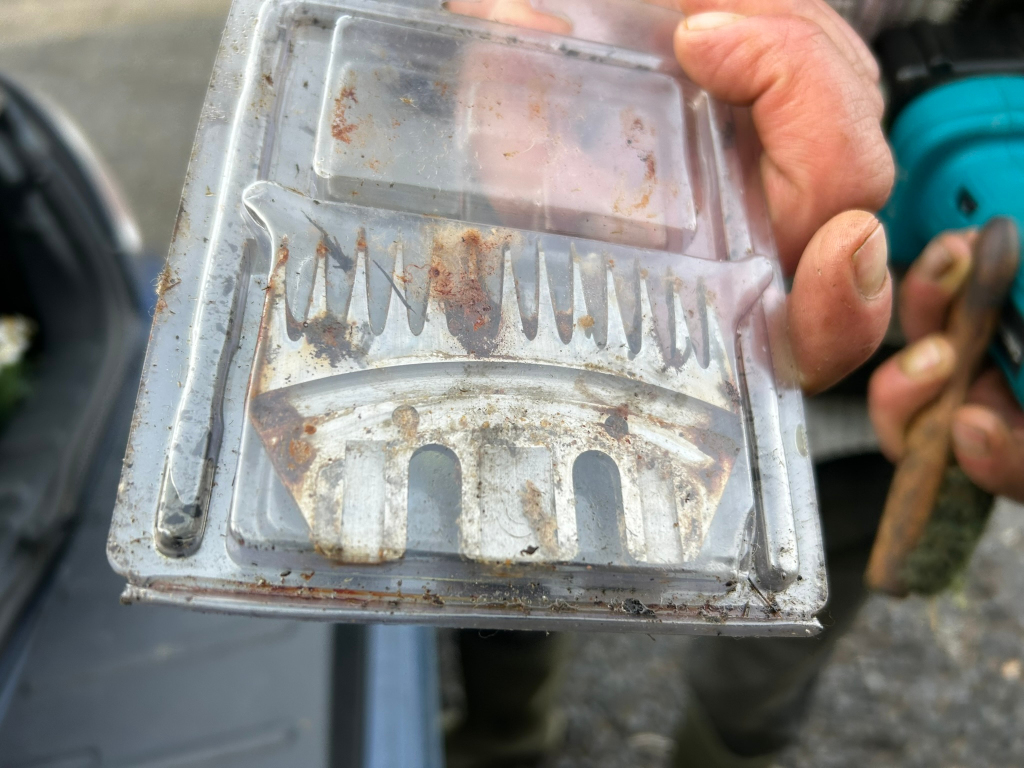 Close-up of a dirty, rusty, and worn-out shearing blade held in a Charlie's hand. The blade is encased in a clear plastic protective casing which is also dirty. The background is blurred, showing part of a vehicle and the person's hand holding what seems to be another tool.