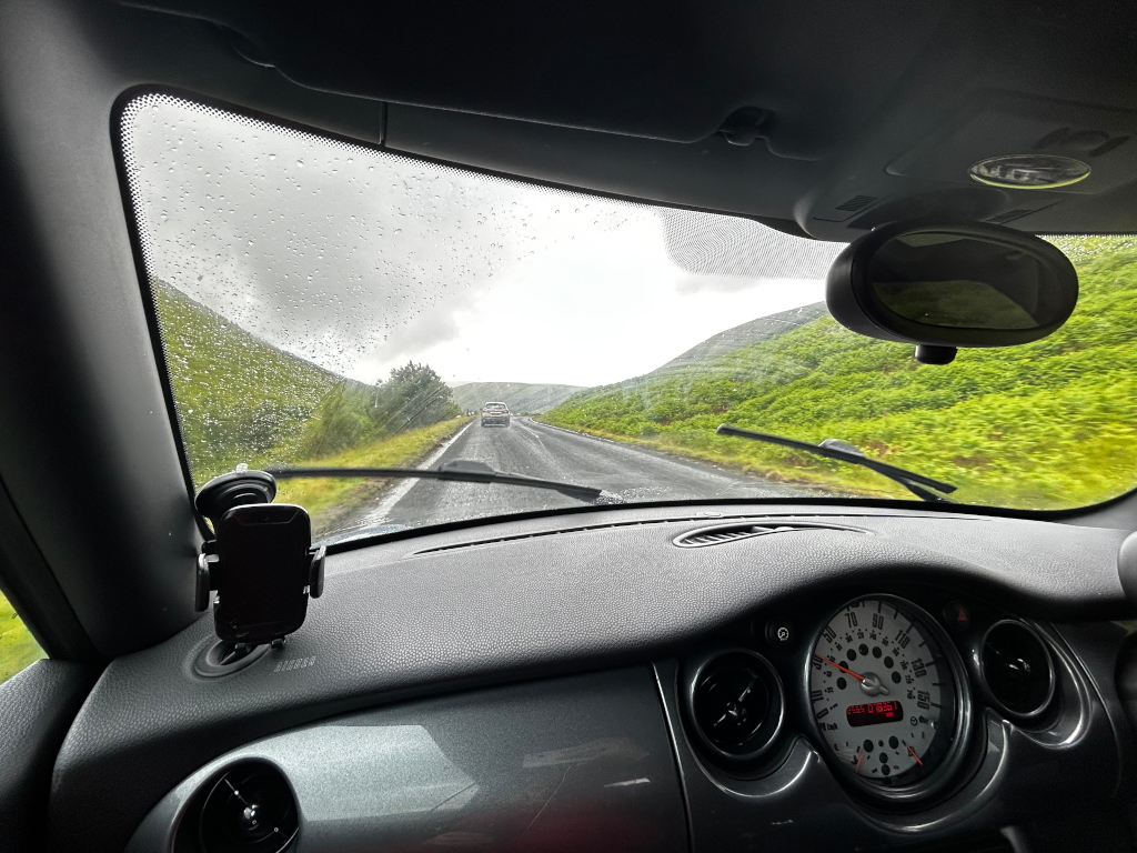View from the inside of a car driving on a wet, winding road. Rain is visible on the windshield, and green hillsides flank the road. Another car is visible in the distance. A phone is mounted on the dashboard. The overall mood is one of journey and possibly slightly melancholic due to the weather.