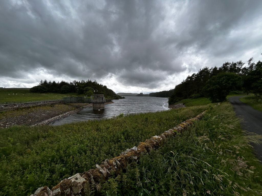 Tranquil scene of a reservoir nestled within a verdant landscape under a brooding, overcast sky. A low stone wall runs along the right, bordering a grassy verge and a paved road. In the middle ground, a small structure, likely a water control mechanism, sits at the edge of the water. The far shore is lined with dark green trees, creating a serene, if somewhat somber, atmosphere.