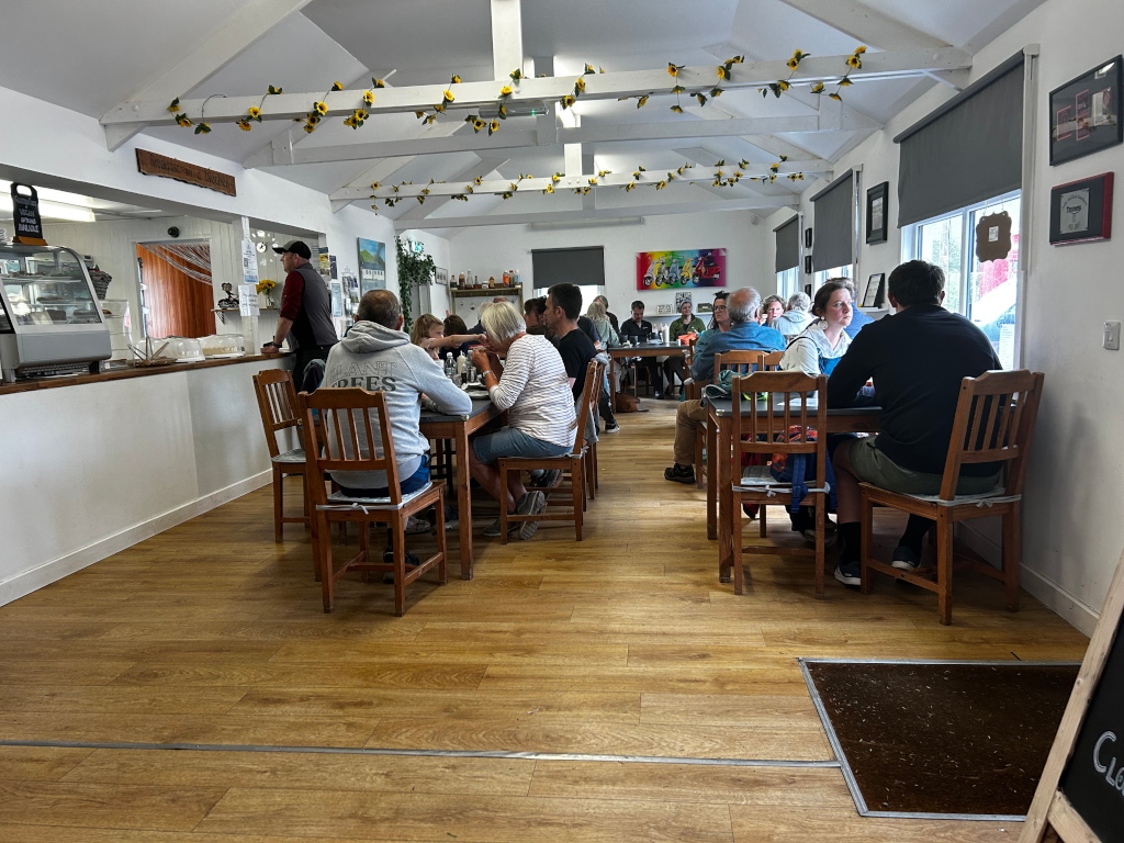 Interior of a cafe or restaurant. Several people are seated at tables, eating and talking. The decor is bright and cheerful, with sunflower decorations hanging from the ceiling. The floor is light brown wood. A counter with a display case is visible at the far end of the room, suggesting a food service area.