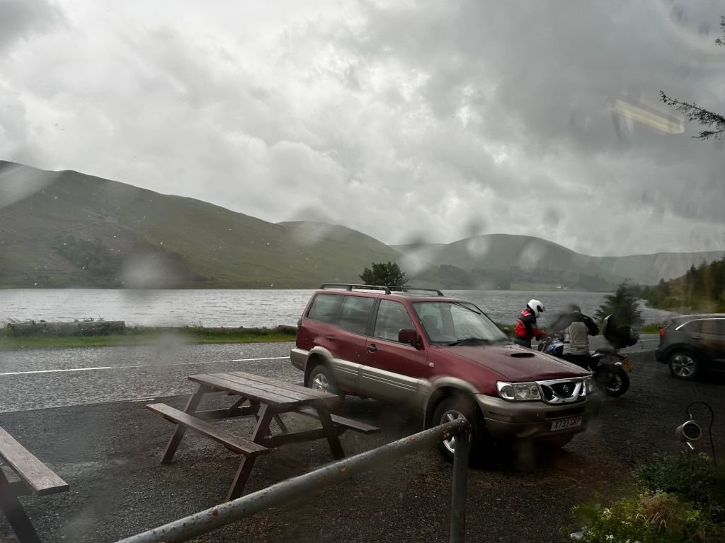 Rainy scene at a lakeside parking area. A maroon SUV is parked next to a picnic table. Two motorcyclists in rain gear are standing near their bikes, which are parked next to the SUV. A dark-coloured car is partially visible in the background. Rolling green hills and a lake are visible in the background. The rain is heavy enough to be seen on the camera lens. The overall mood is sombre and slightly melancholic, due to the weather and the somewhat deserted setting.