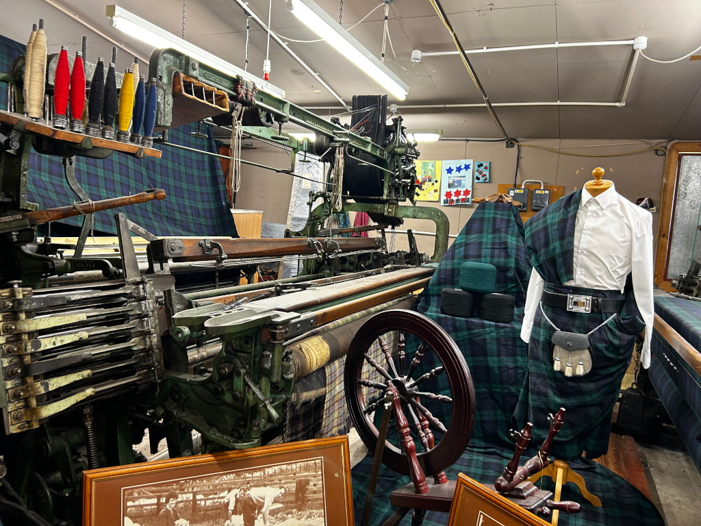 Vintage loom in a workshop, possibly a tartan weaving mill. The loom is predominantly green and features a collection of colourful bobbins. A partially draped kilt and a mannequin dressed in a white shirt and kilt are displayed in front of the loom, suggesting the production and display of the finished product. Framed photos or documents are also visible on the floor, likely showcasing the workshop's history or product. The overall scene conveys a sense of traditional craftsmanship and textile production.