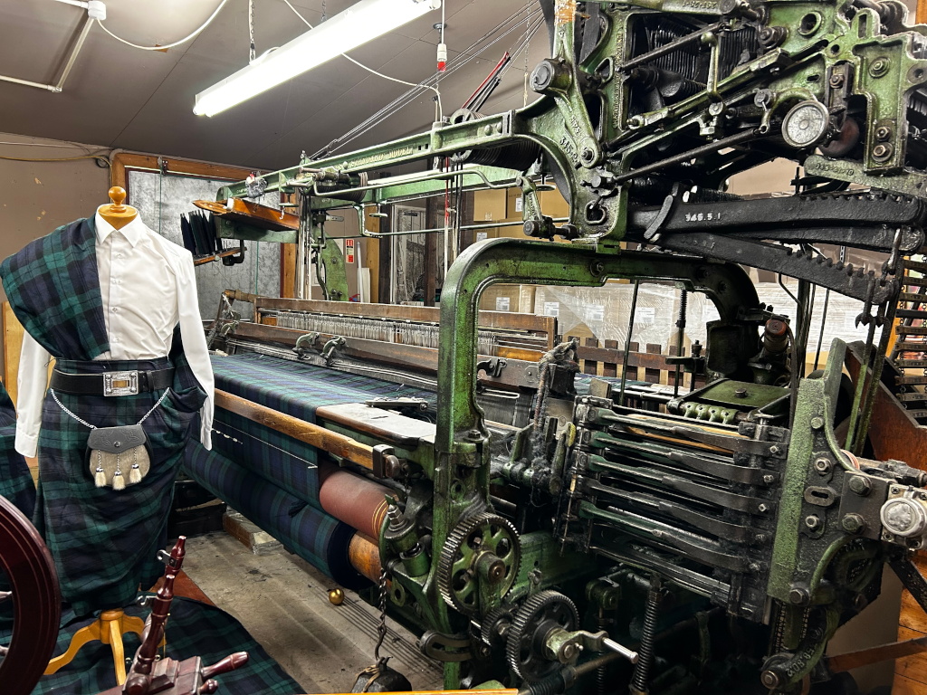 Large, antique-looking green loom, prominently featured in the centre. A partially completed tartan fabric is visible on the loom, its dark green and black pattern clearly displayed. Next to the loom, a mannequin is dressed in a matching tartan kilt, sporran, and white shirt, serving as a display of the finished product.