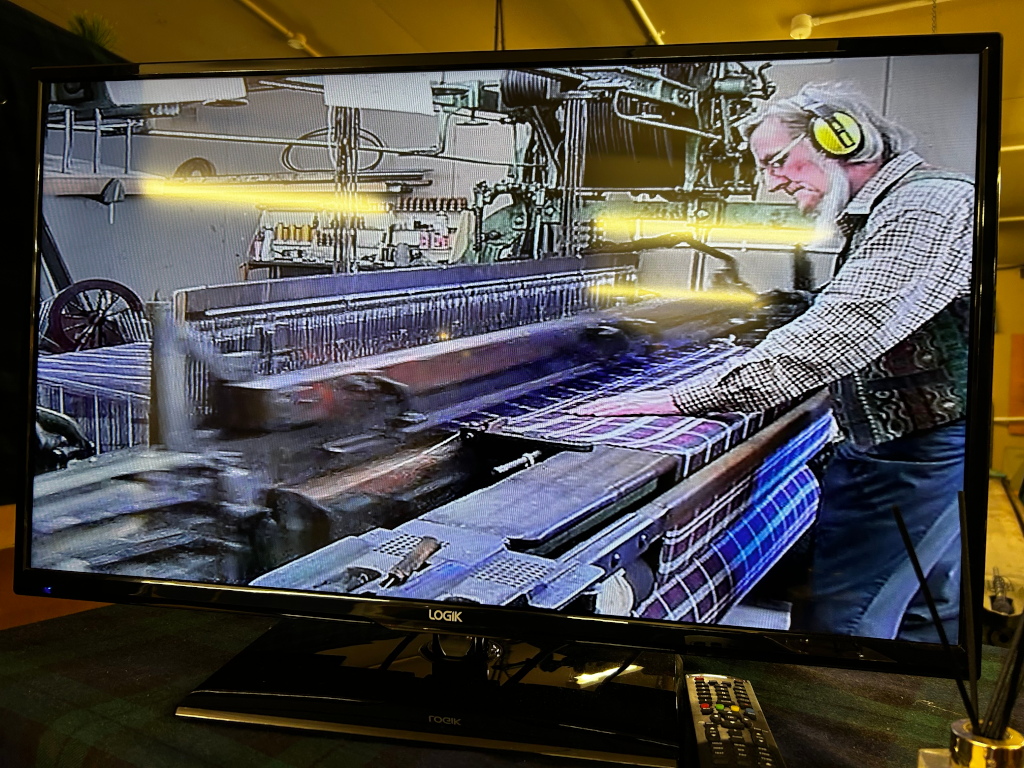Flat-screen television displaying a video of a man operating a traditional loom, weaving plaid fabric. The video appears to be of older recording quality. The man is wearing hearing protection and is focused on his work. The overall scene depicts a process of traditional textile manufacturing.