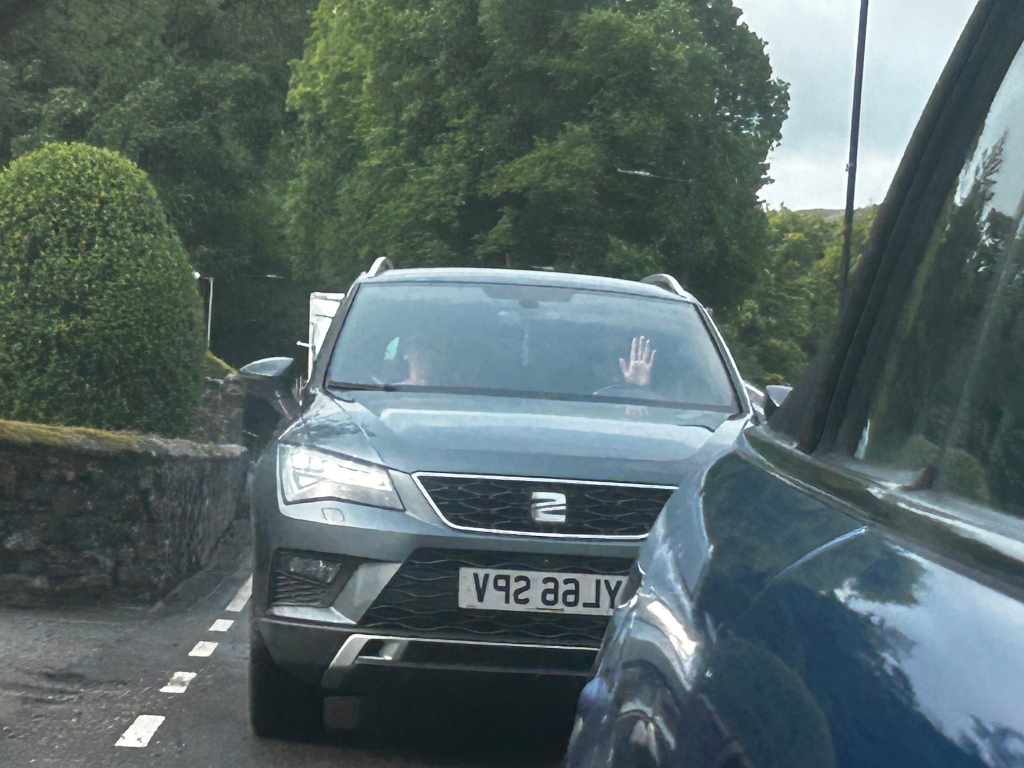 Grey Seat Ateca SUV stopped on a narrow road, partially obscured by another vehicle. A person inside the Seat is hold the palm of their hand against the window. The setting appears to be rural, with lush green foliage and a stone wall visible. The focus is on the Seat and the hand, creating a sense of a brief, possibly fleeting, interaction between the occupants of the two vehicles.