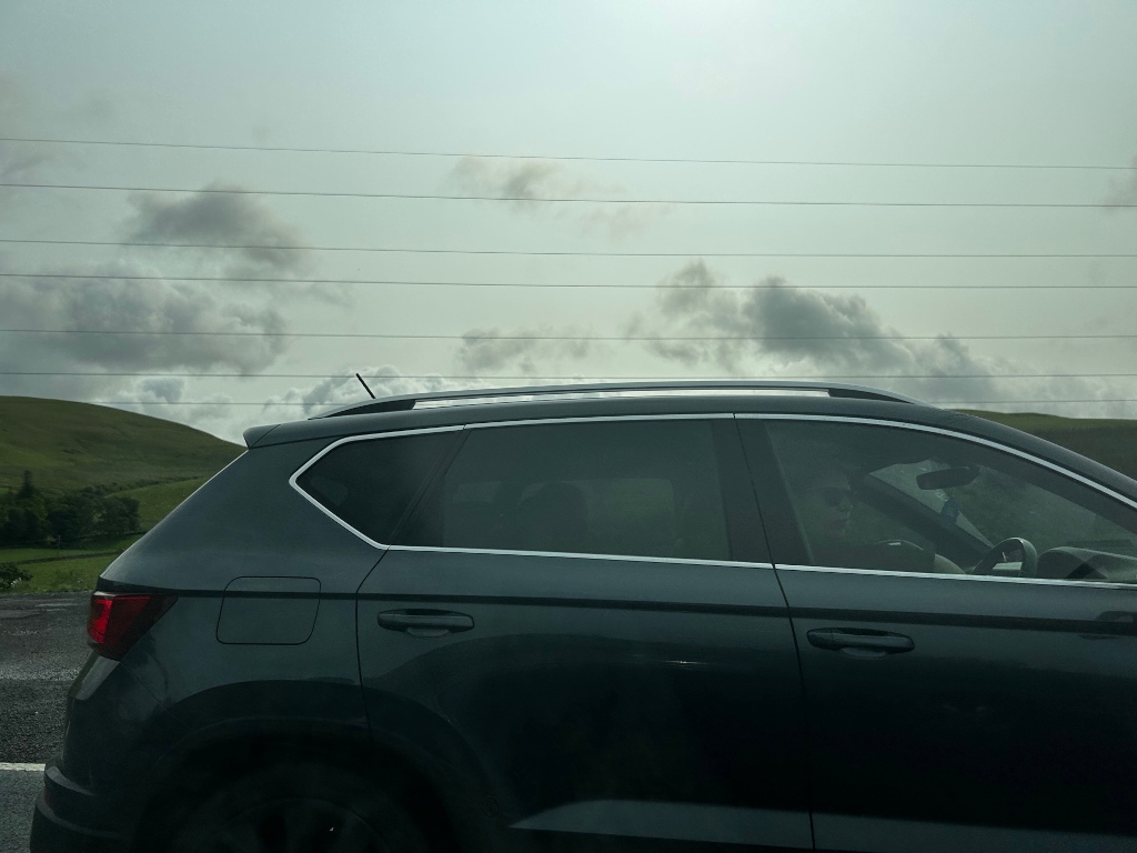 Side of a dark-coloured SUV driving along a road. The vehicle is in the foreground, with a view of rolling green hills and a cloudy sky in the background. Power lines stretch across the image above the hills.