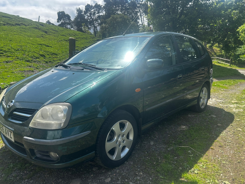 Dark green Nissan Almeria parked on a gravel drive. The car appears to be in relatively good condition, and the background features a grassy hillside and some trees. The overall impression is a simple, everyday scene of a car parked in a rural setting. There's nothing particularly striking or symbolic about the picture; it's a straightforward depiction of a vehicle in a landscape.