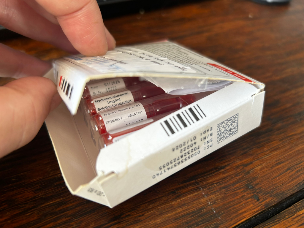 Leonie's hand opening a box of Hydroxocobalamin 1mg/ml injection vials. The box is white and clearly labelled with the drug's name, concentration, manufacturer (Accord Healthcare Ltd), lot number, and expiry date. The vials themselves are visible inside the packaging. The overall impression is one of medical treatment or preparation for an injection.