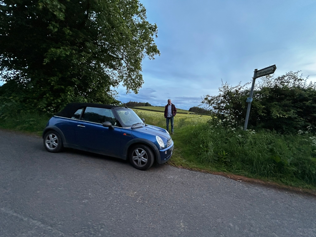 Country road lined with tall trees on either side. The road is relatively empty and the light suggests it is either early morning or late evening. The trees are lush and green, and the overall atmosphere is tranquil and serene. The road leads toward a slightly visible horizon with fields beyond the trees.