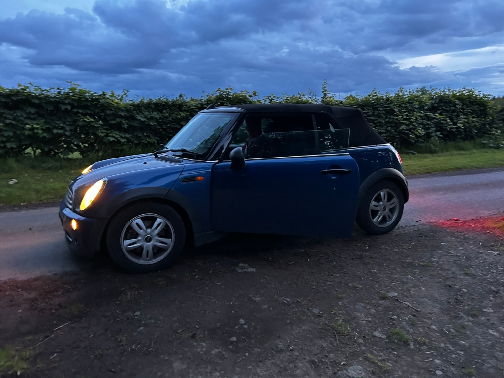 Blue Mini Cooper convertible parked on the side of a country road at dusk. The sky is cloudy, and a hedge runs along the roadside. The car's headlights are on, suggesting it's either early evening or late in the day. The overall mood is peaceful and somewhat melancholic, due to the subdued lighting and the quiet rural setting.