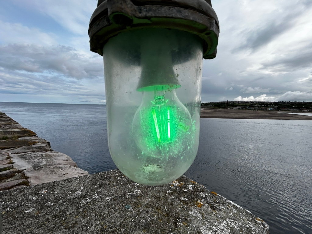 Close-up view of a weathered, cylindrical glass lamp fixture containing a glowing green light bulb. The lamp is affixed to a stone structure overlooking a body of water, possibly a river or sea, with a distant shoreline visible under a cloudy sky. The overall mood is somewhat melancholic or atmospheric, due to the muted colours and the slightly ominous weather.