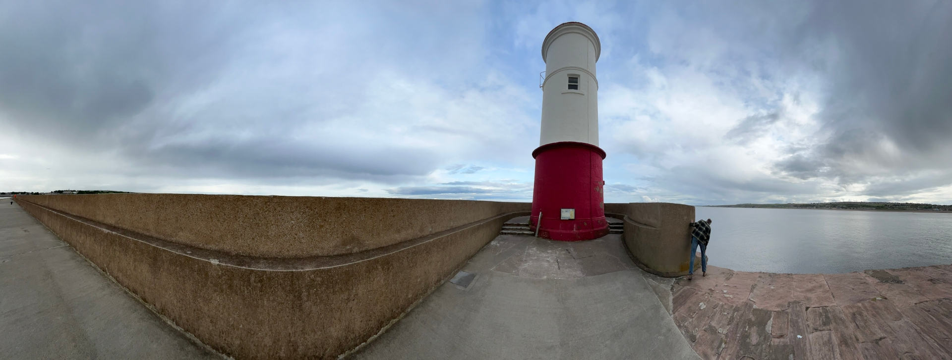 Panoramic view of a lighthouse situated on a concrete pier overlooking a calm body of water. The sky is overcast with a mix of grey and white clouds. A person is visible in the lower right corner, seemingly observing the scene. The lighthouse is predominantly white with a red base, and the pier features a textured concrete wall.